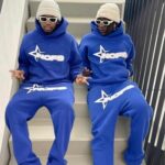 Two individuals wearing matching blue NOFS tracksuits with bold white branding, white sunglasses, and beige beanies, sitting on a staircase with a stylish urban aesthetic.