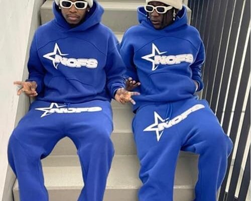 Two individuals wearing matching blue NOFS tracksuits with bold white branding, white sunglasses, and beige beanies, sitting on a staircase with a stylish urban aesthetic.