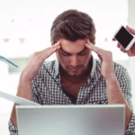 Stressed man at desk surrounded by documents, phone, and pencil, showing stress and pressure at work.